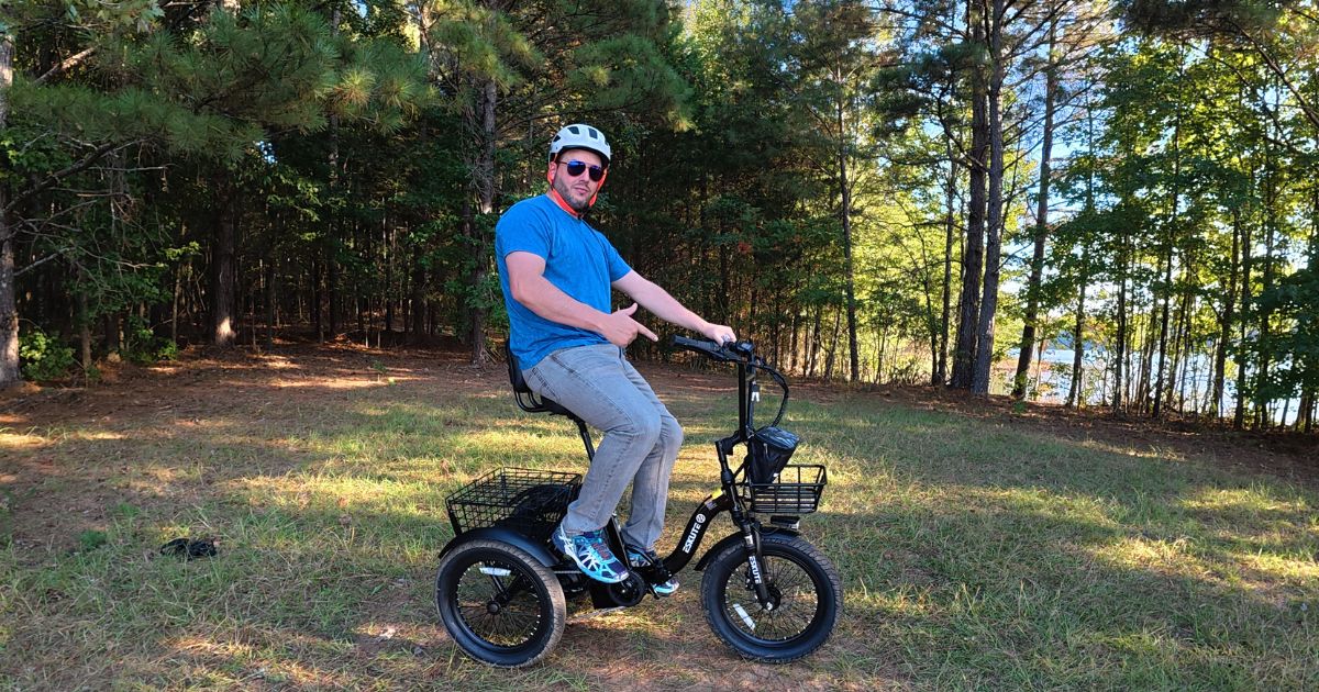 A man wearing a helmet and sunglasses sitting on a black electric tricycle in a wooded area, pointing forward