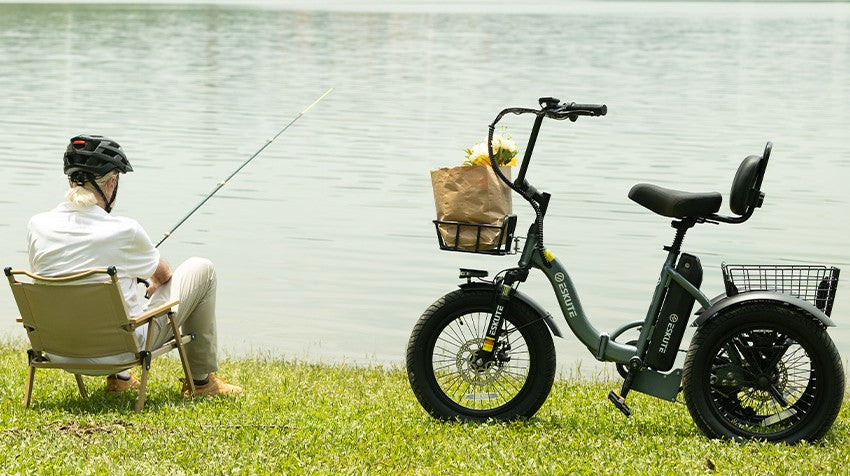 An older man wearing a helmet sitting in a folding chair fishing by a lake, with a grey Eskute electric tricycle parked on the grass beside him