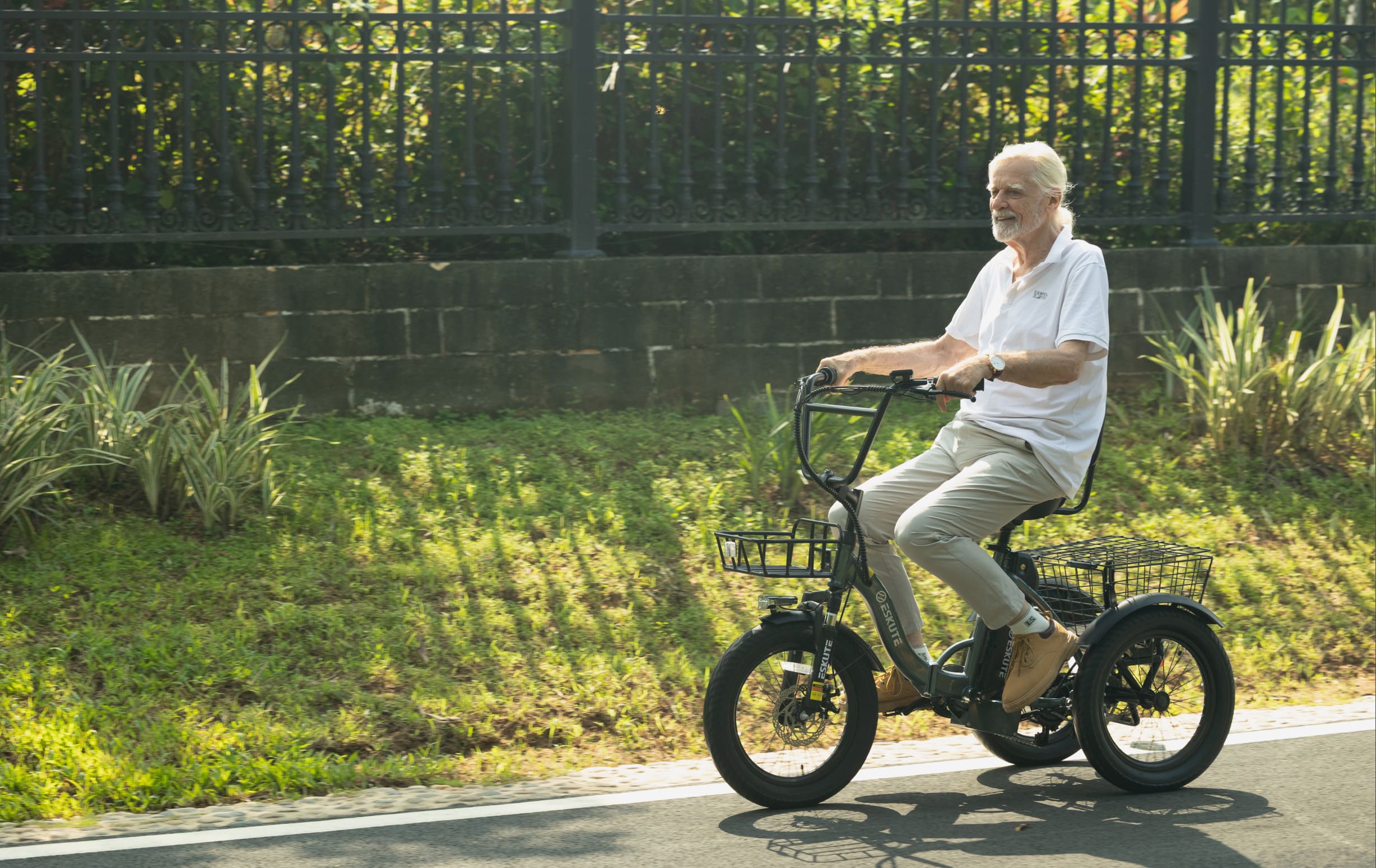 Senior man riding a black Eskute three-wheel electric trike along a sunny tree-lined road, with front and rear cargo baskets