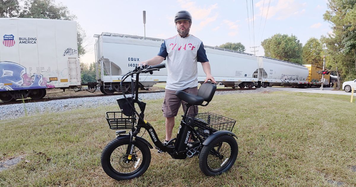 man wearing a helmet standing next to a black ESKUTE three-wheeled electric bike, with freight train cars and railway tracks in the background